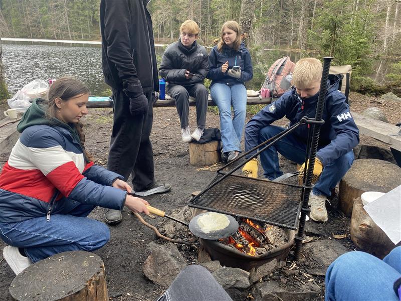 Gruppe von Personen am Lagerfeuer, einer kocht über dem Feuer. Wald und See im Hintergrund.