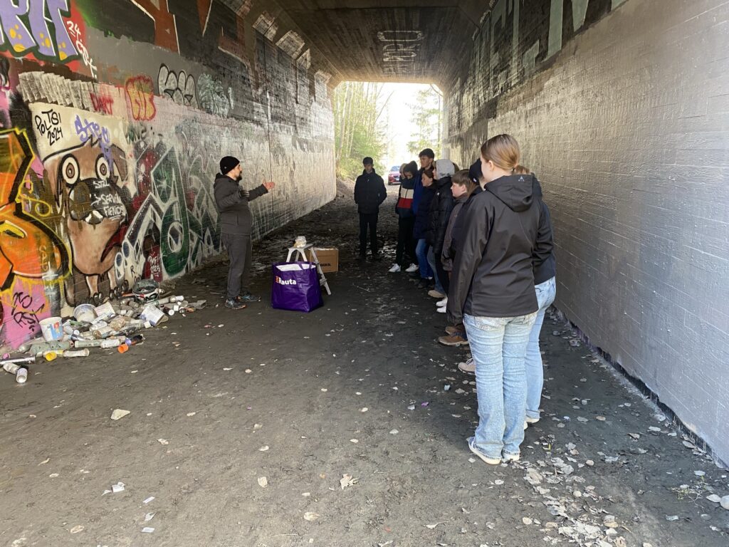 Gruppe von Menschen hört einer Person unter einer Brücke zu, umgeben von Graffiti.