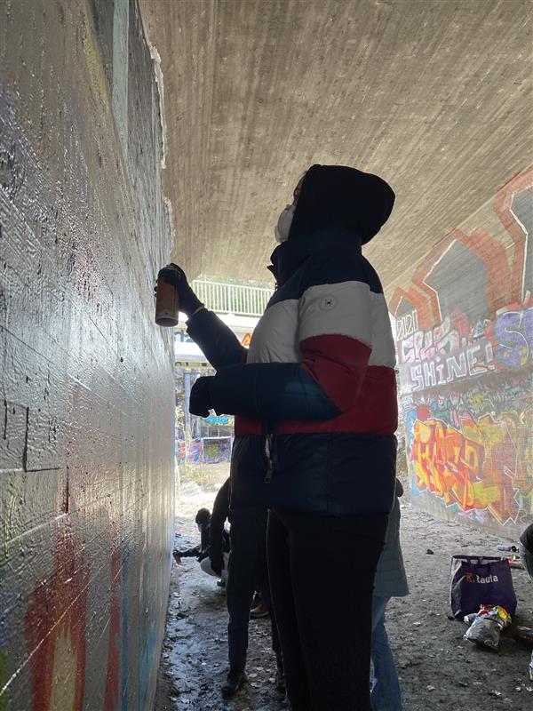 Ein Person sprayt Farbe an eine Wand unter einer Brücke.