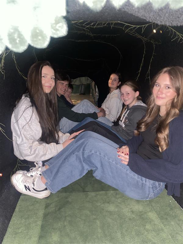 Schüler sitzen in einem dunkel gestalteten Raum auf einer grünen Fläche.