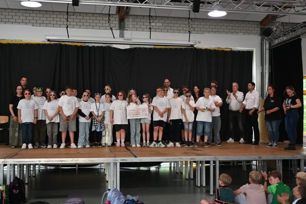 Schülergruppe auf der Bühne, lächelnd, in weißen T-Shirts und Sonnenbrillen.