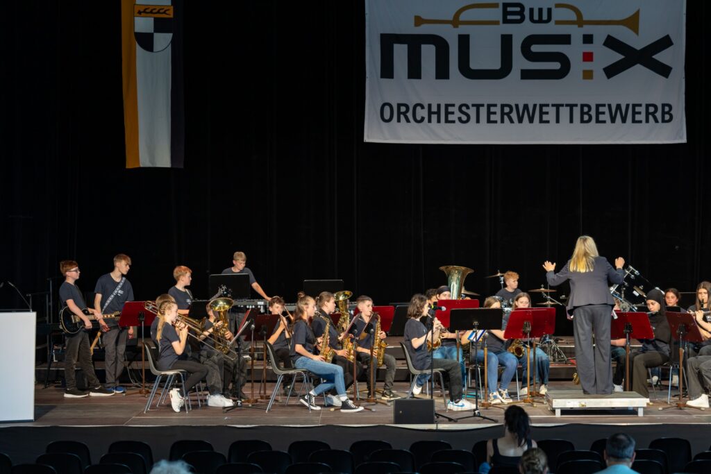 Gesamtaufnahme der Bühne mit der Bläserklasse 7d beim BW Musix-Wettbewerb – das gesamte Orchester ist in Aktion, begleitet von intensiver Dirigierarbeit durch Christine Jahn.