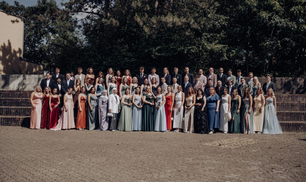 Gruppenfoto des Abiturjahrgangs 2025 der Gesamtschule Waldbröl auf dem Schulhof – die Abiturient:innen stehen festlich gekleidet in mehreren Reihen vor einer grünen Baumkulisse und lächeln in die Kamera.