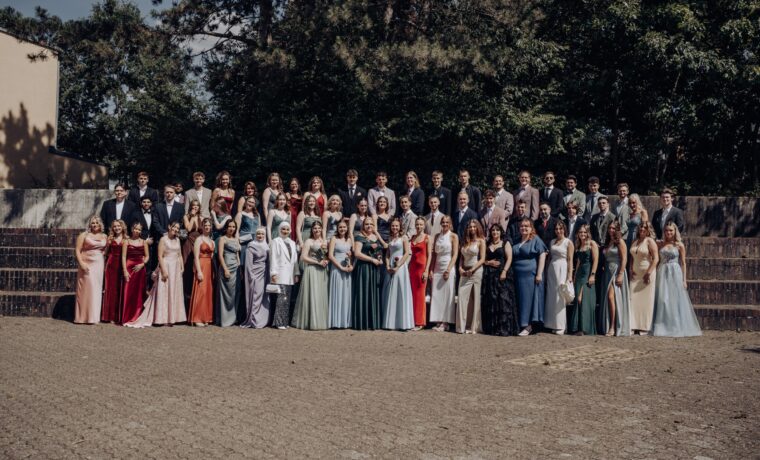 Gruppenfoto des Abiturjahrgangs 2025 der Gesamtschule Waldbröl auf dem Schulhof – die Abiturient:innen stehen festlich gekleidet in mehreren Reihen vor einer grünen Baumkulisse und lächeln in die Kamera.