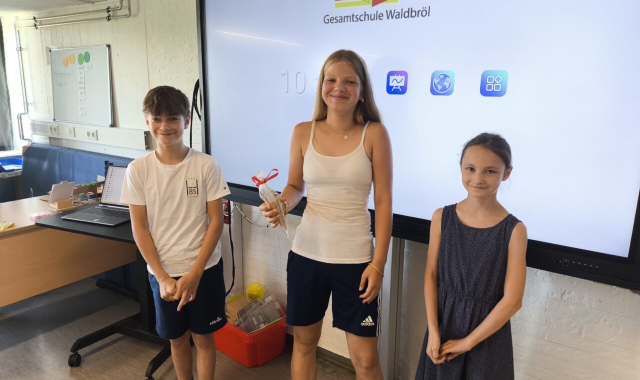 Drei strahlende Schüler:innen stehen vor einem digitalen Whiteboard mit dem Logo der Gesamtschule Waldbröl. In der Mitte hält eine Schülerin den Eisgutschein in der Hand, überreicht von den beiden SV-Initiatorinnen des Fahrradwettbewerbs.