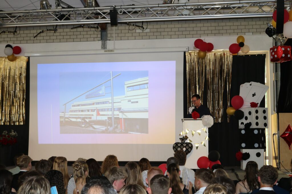 Paul Giebeler bei seinem spontanen Grußwort am Pult, im Hintergrund ein Foto der Schule auf der Leinwand, davor das gespannte Publikum.
