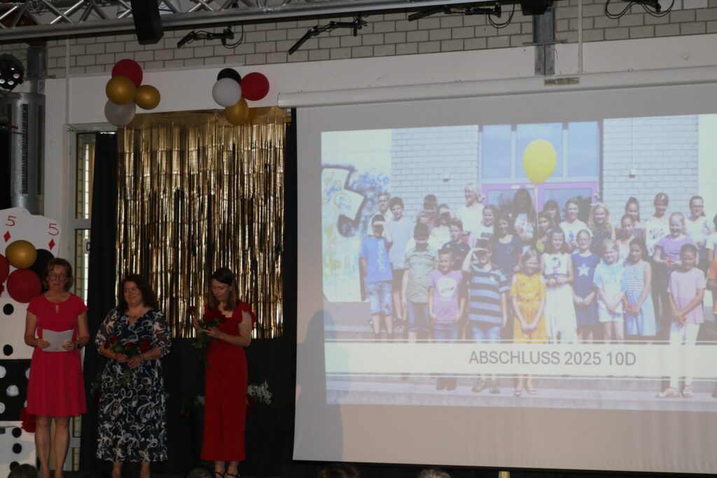 Drei Lehrerinnen präsentieren die Abschlussklasse 10d, während im Hintergrund ein altes Klassenfoto auf der Leinwand erscheint.