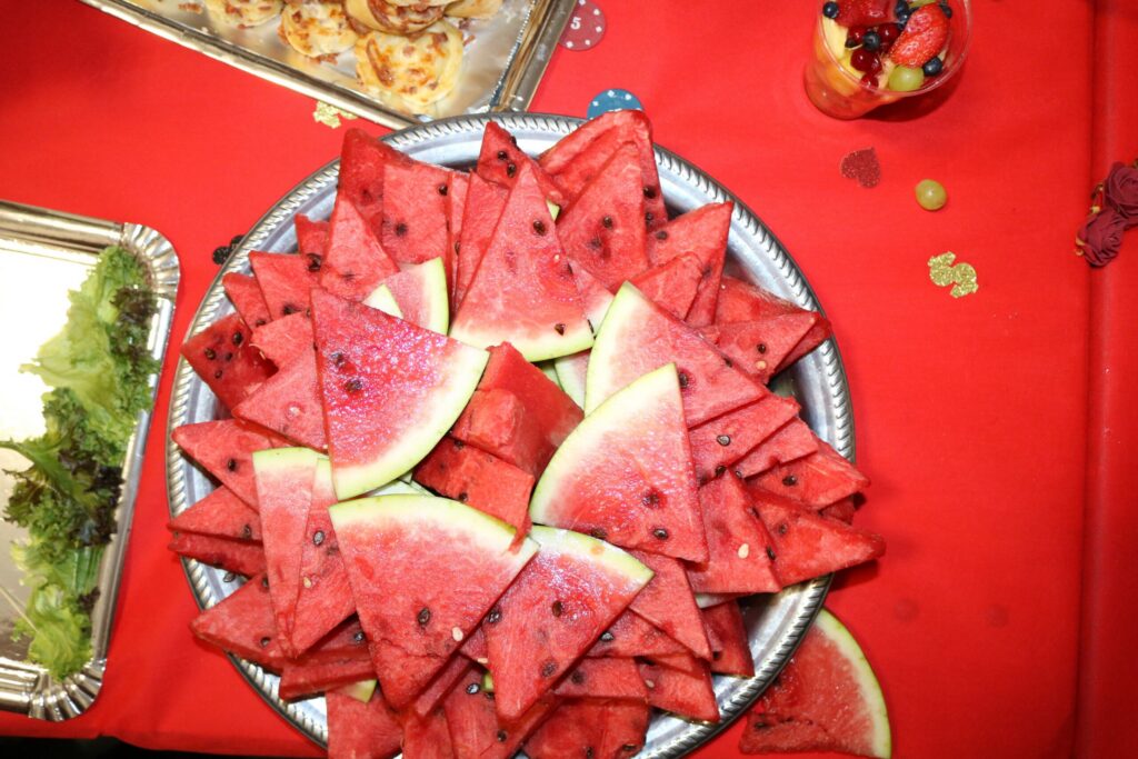 Frische Wassermelone in Dreiecks-Stücken auf einem silbernen Tablett – sommerlich angerichtet, umgeben von rotem Tischtuch und Dekokonfetti in Pokerchips-Optik.