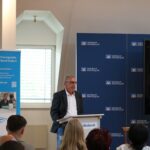 Grußwort durch einen Vertreter des Schulamts auf der Bühne der Volksbank Oberberg. Im Hintergrund ein Roll-Up der kommunalen Koordinierungsstelle.