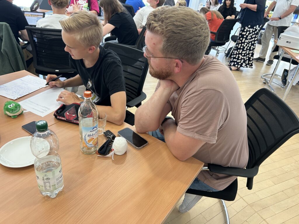 Ein Jugendlicher schreibt Notizen auf ein Arbeitsblatt, neben ihm sitzt ein Betreuer; auf dem Tisch stehen Getränke, Smartphones und kleine Maskottchen.
