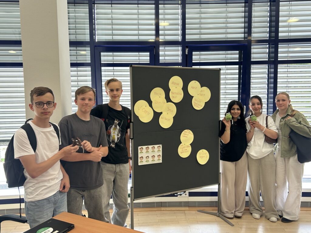 Die Gruppe der Social Media Scouts steht vor einer Präsentationstafel mit Stichworten zum Thema Medienkompetenz; sie halten kleine Maskottchen in den Händen.
