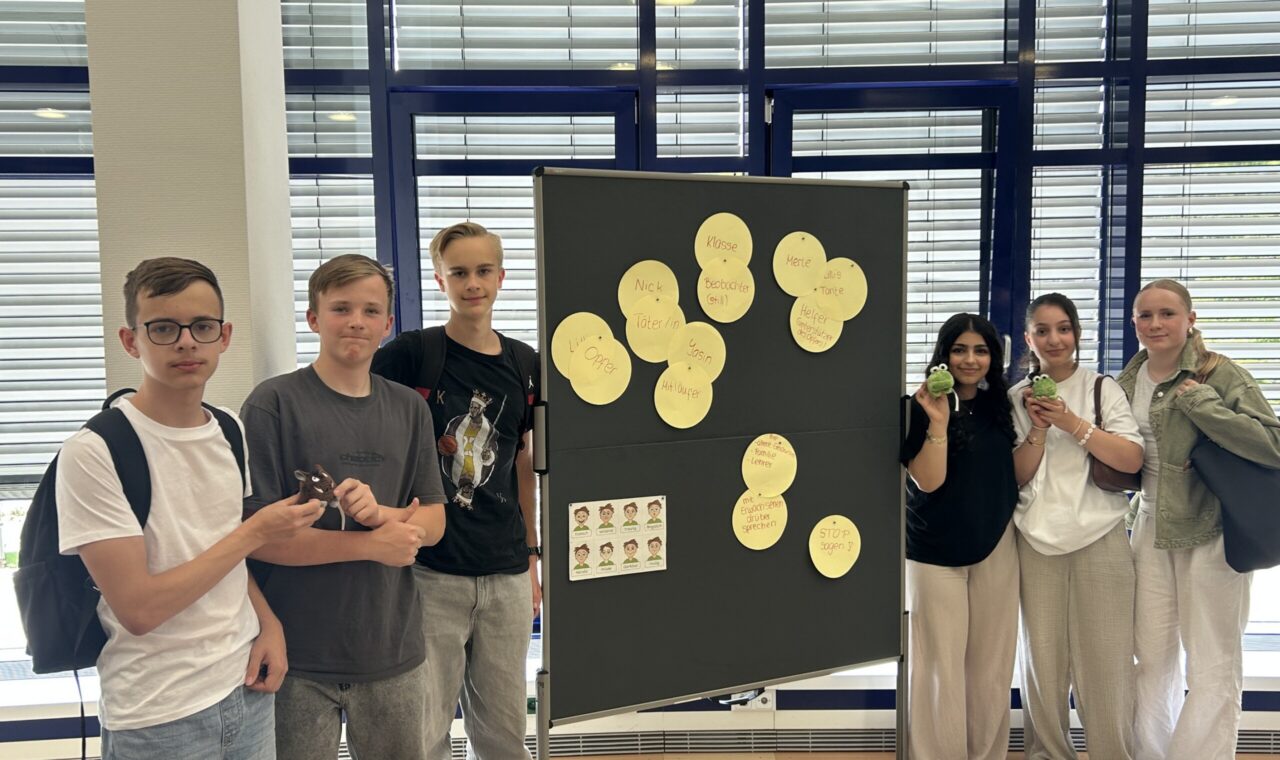 Die Gruppe der Social Media Scouts steht vor einer Präsentationstafel mit Stichworten zum Thema Medienkompetenz; sie halten kleine Maskottchen in den Händen.