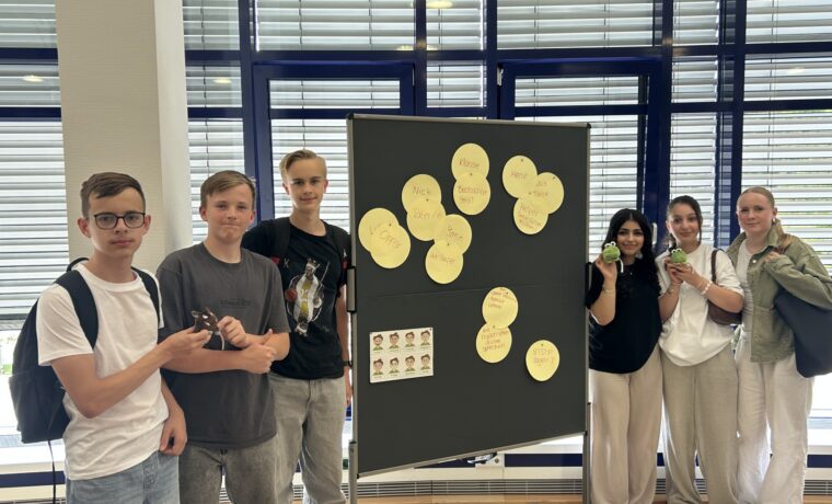 Die Gruppe der Social Media Scouts steht vor einer Präsentationstafel mit Stichworten zum Thema Medienkompetenz; sie halten kleine Maskottchen in den Händen.