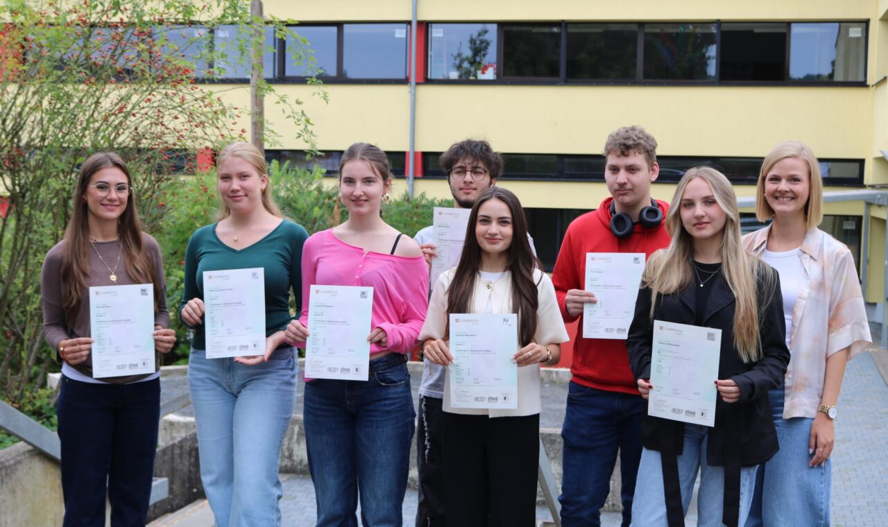 Acht Schüler:innen des Cambridge-Projektkurses der Gesamtschule Waldbröl halten stolz ihre Cambridge-Zertifikate in den Händen, gemeinsam mit Kursleiterin Lisa Dornseifer.