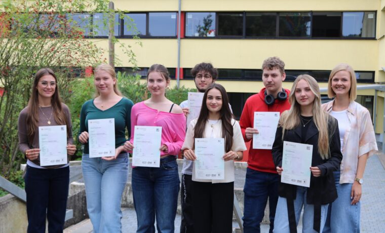 Acht Schüler:innen des Cambridge-Projektkurses der Gesamtschule Waldbröl halten stolz ihre Cambridge-Zertifikate in den Händen, gemeinsam mit Kursleiterin Lisa Dornseifer.