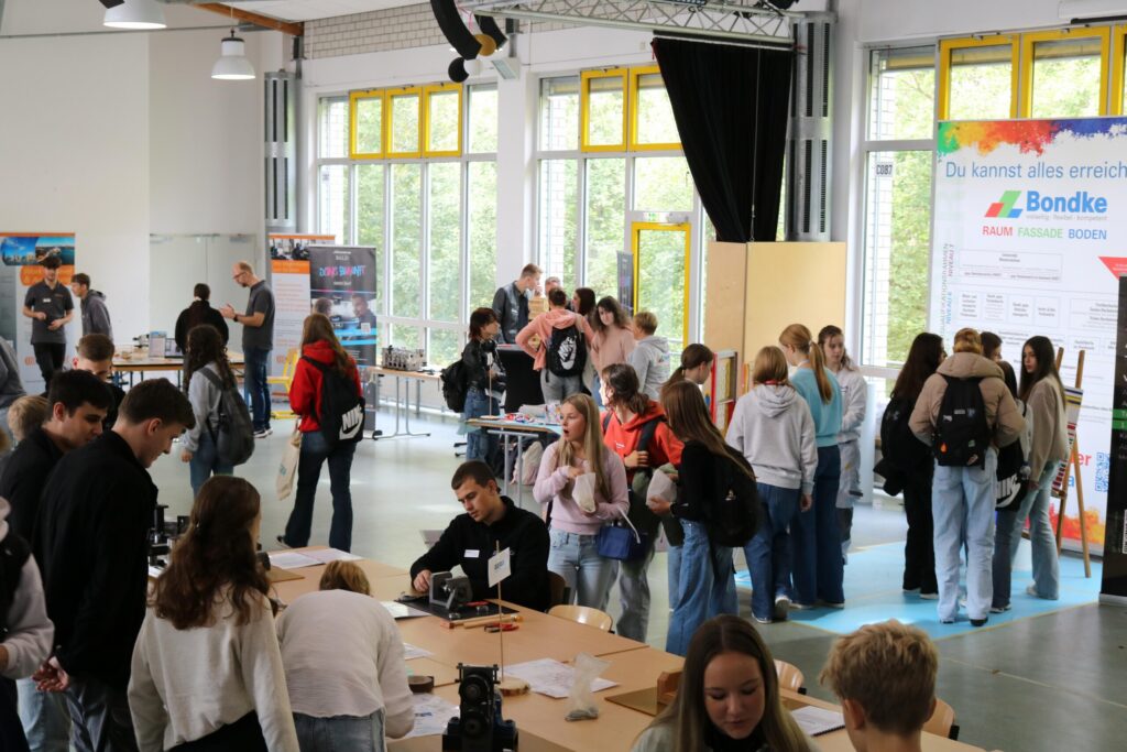 Schülerinnen und Schüler stehen in einer Schulmensa an verschiedenen Ständen, informieren sich und führen Gespräche.