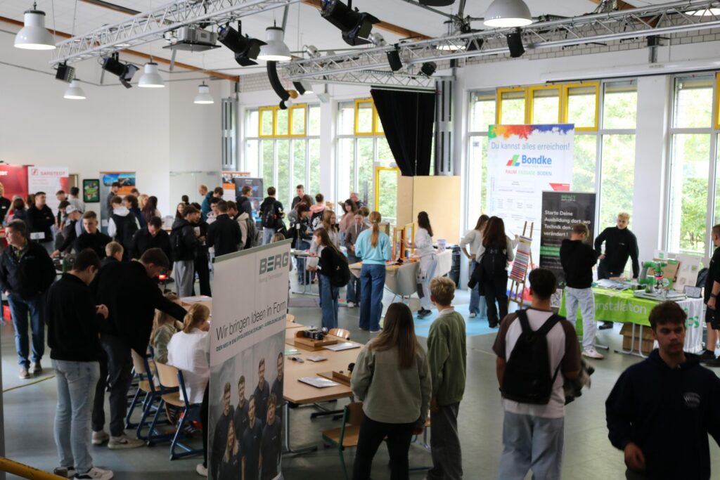 Viele Schülerinnen und Schüler bewegen sich durch einen großen Raum mit zahlreichen Infoständen. Gespräche und Begegnungen prägen die Atmosphäre.