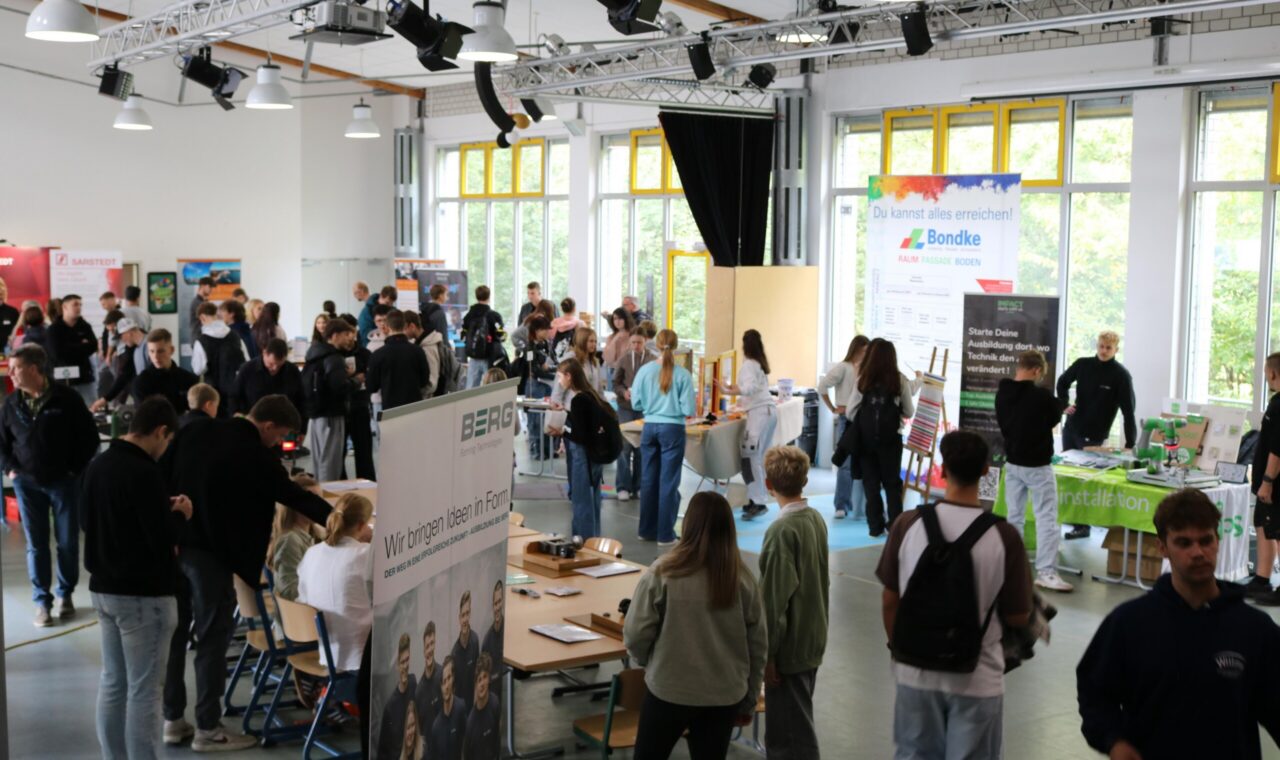 Viele Schülerinnen und Schüler bewegen sich durch einen großen Raum mit zahlreichen Infoständen. Gespräche und Begegnungen prägen die Atmosphäre.