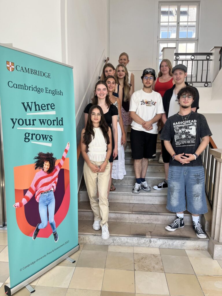 Die Teilnehmer:innen des Cambridge-Projektkurses stehen gemeinsam mit Lisa Dornseifer im Englischen Institut in Köln an einem Cambridge-Banner auf einer Treppe.