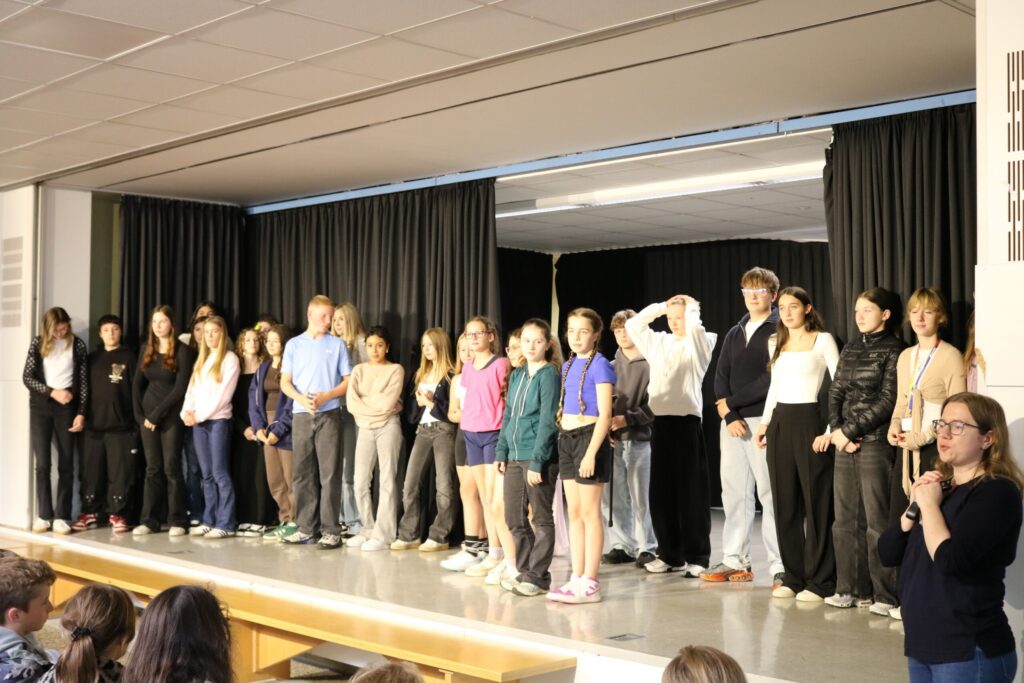 Alle Mitwirkenden der Open Stage – Schülerinnen und Schüler verschiedener Klassen sowie der DG-Kurs 9 – stehen gemeinsam auf der Bühne. Sie bilden eine lange Reihe und lassen sich vom Publikum feiern. Am rechten Bühnenrand ist eine Lehrkraft mit Mikrofon zu sehen, die die Veranstaltung moderiert.