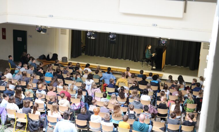 Anne Mauracher aus der Klasse 6d sitzt am Klavier und spielt die Stücke „Zerflossene Melodie“ und „Tanz der Intervalle“. Vor ihr füllt sich das Forum mit den 5. Klassen, die dicht gedrängt auf Stühlen sitzen und gespannt zuhören. Das Foto zeigt die gesamte Probebühne im architektonischen Rahmen des Forums.