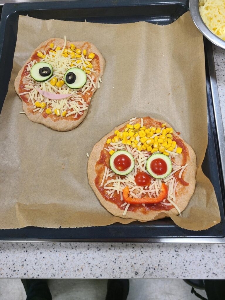 Zwei kleine selbstgemachte Pizzen auf einem Backblech, liebevoll mit Gemüse zu lachenden Gesichtern dekoriert.