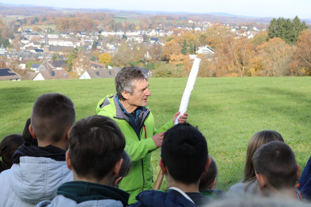 „Johannes Heibel steht mit einer zusammengerollten Weißen Friedensfahne vor einer Gruppe von Schüler:innen auf einer Wiese oberhalb von Waldbröl. Im Hintergrund ist die herbstliche Stadtlandschaft zu sehen.“