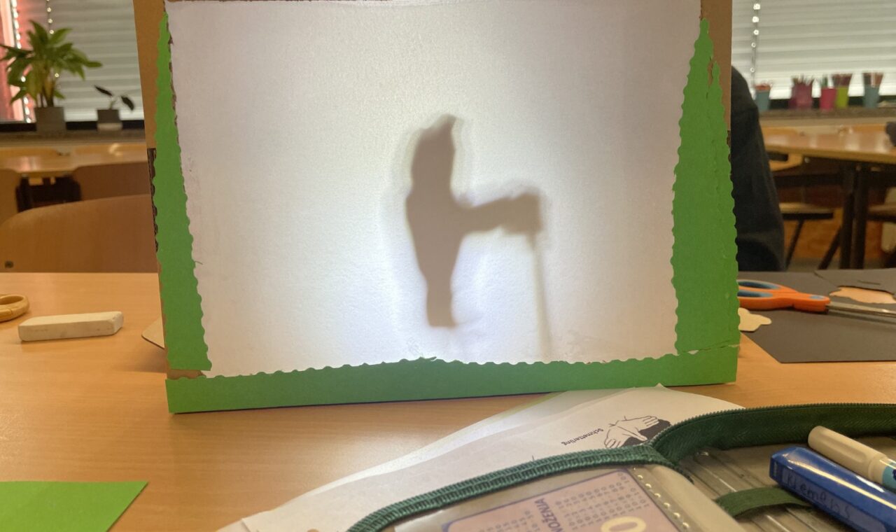 Ein selbstgebautes Schattentheater steht auf einem Tisch, dahinter ist die Silhouette einer Figur zu erkennen.