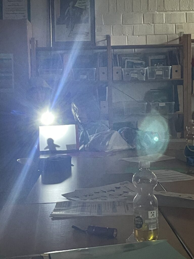 Ein heller Lichtstrahl eines Projektors fällt in einem dunklen Raum auf eine kleine Schattenbühne, auf der die Silhouette einer Figur zu sehen ist.
