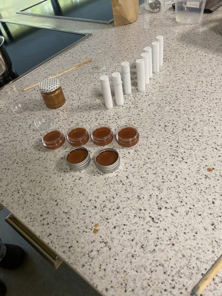 Mehrere geöffnete Kosmetiktiegel mit brauner Creme stehen auf einer Arbeitsfläche, daneben leere Lippenpflegestifthüllen.