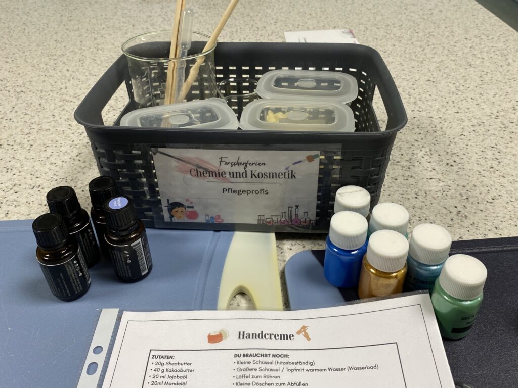 Materialien für den Workshop „Chemie und Kosmetik“, darunter Bechergläser, ätherische Öle, Farbpigmente und ein Rezept für Handcreme.
