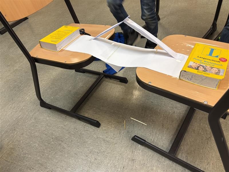 Zwei Stühle, dazwischen eine Papierbrücke, die mit Büchern beschwert ist – ein Technik-Experiment im Klassenzimmer.