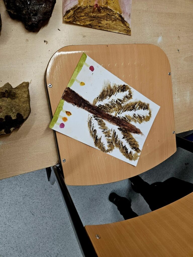 Ein Kinderbild mit einem Baum, gemalt in Naturfarben, liegt auf einem Stuhl neben weiteren Werkstücken.