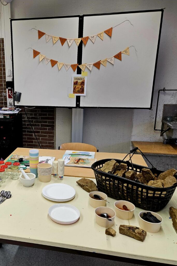 Ein vorbereiteter Stationstisch zur „Steinzeit“-Werkstatt mit Naturmaterialien, Schalen, Steinen und Arbeitsutensilien vor einem Whiteboard mit der Aufschrift „Willkommen Höhlenforscher“.