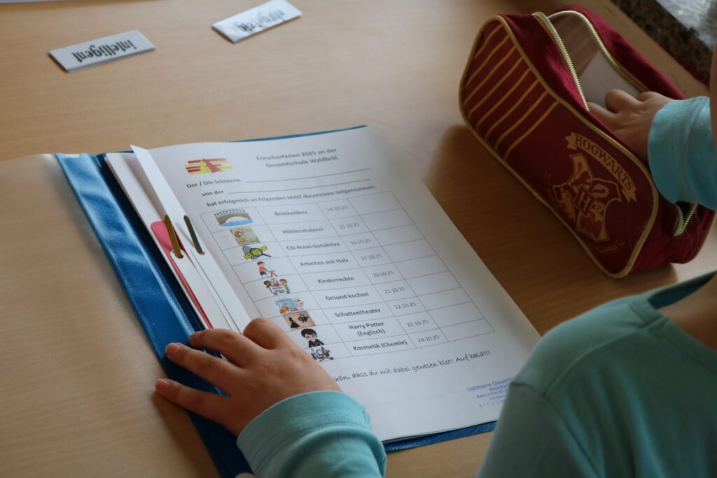 Ein Kind schaut auf seine gefüllte Forscherferien-Mappe mit eingetragenen absolvierten Workshops, daneben ein Mäppchen mit Hogwarts-Motiv.