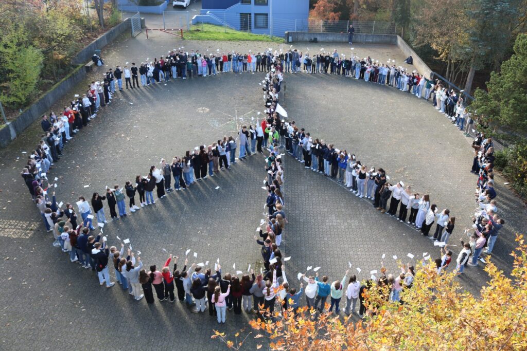 „Rund 300 Schüler:innen stehen auf dem Schulhof der Gesamtschule Waldbröl in einer großen Formation und bilden ein Peace-Zeichen. Viele halten kleine weiße Fahnen in die Höhe.“