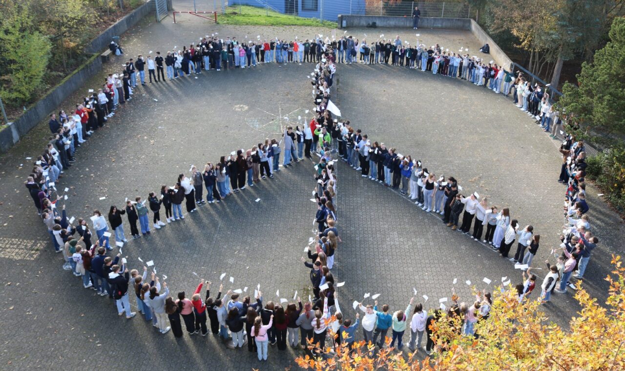 „Rund 300 Schüler:innen stehen auf dem Schulhof der Gesamtschule Waldbröl in einer großen Formation und bilden ein Peace-Zeichen. Viele halten kleine weiße Fahnen in die Höhe.“