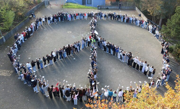 „Rund 300 Schüler:innen stehen auf dem Schulhof der Gesamtschule Waldbröl in einer großen Formation und bilden ein Peace-Zeichen. Viele halten kleine weiße Fahnen in die Höhe.“