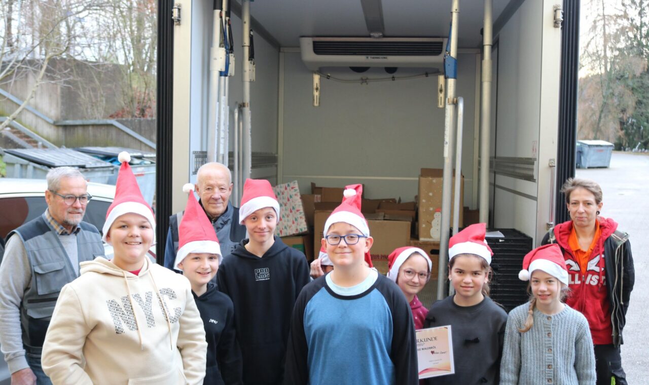 Eine Gruppe von neun Schüler:innen mit Weihnachtsmützen steht vor einem geöffneten Lieferwagen der Tafel Oberberg Süd. Hinter ihnen sind mehrere Kartons sichtbar. Zwei erwachsene Mitarbeitende der Tafel sowie eine erwachsene Begleitperson stehen am Rand. Ein Kind hält eine Urkunde in der Hand.