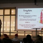 Moderatorin steht auf der Bühne und erklärt den Ablauf des Vorlesewettbewerbs vor einer projizierten Folie mit dem Titel „Vorgehen“.