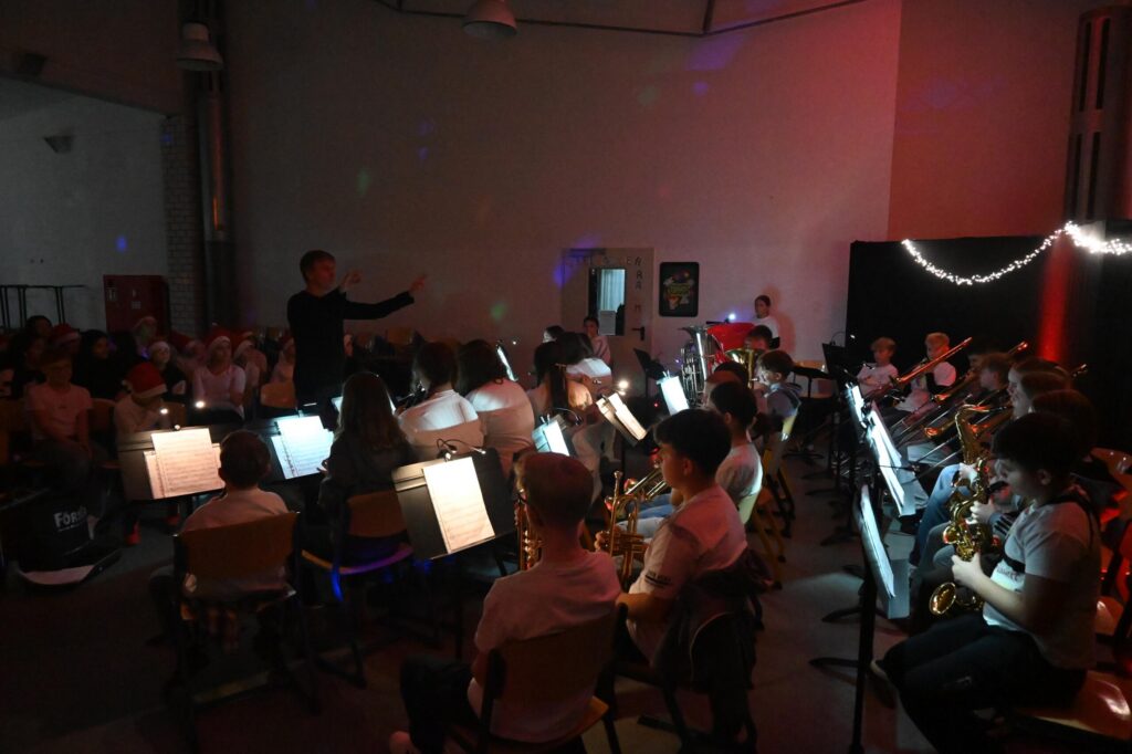 Bläserklasse der Gesamtschule sitzt halbrund auf der Bühne, dirigiert von einem Lehrer, mit Notenständern und Blasinstrumenten, stimmungsvolle Beleuchtung während der Veranstaltung „Lichter im Advent“.