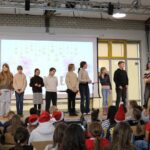Acht Schüler:innen stehen nebeneinander auf der Bühne vor einer Leinwand mit der „Finale“-Folie; eine Moderatorin und ein Jurymitglied sprechen ins Mikrofon, das Publikum sitzt aufmerksam davor in der Mensa.