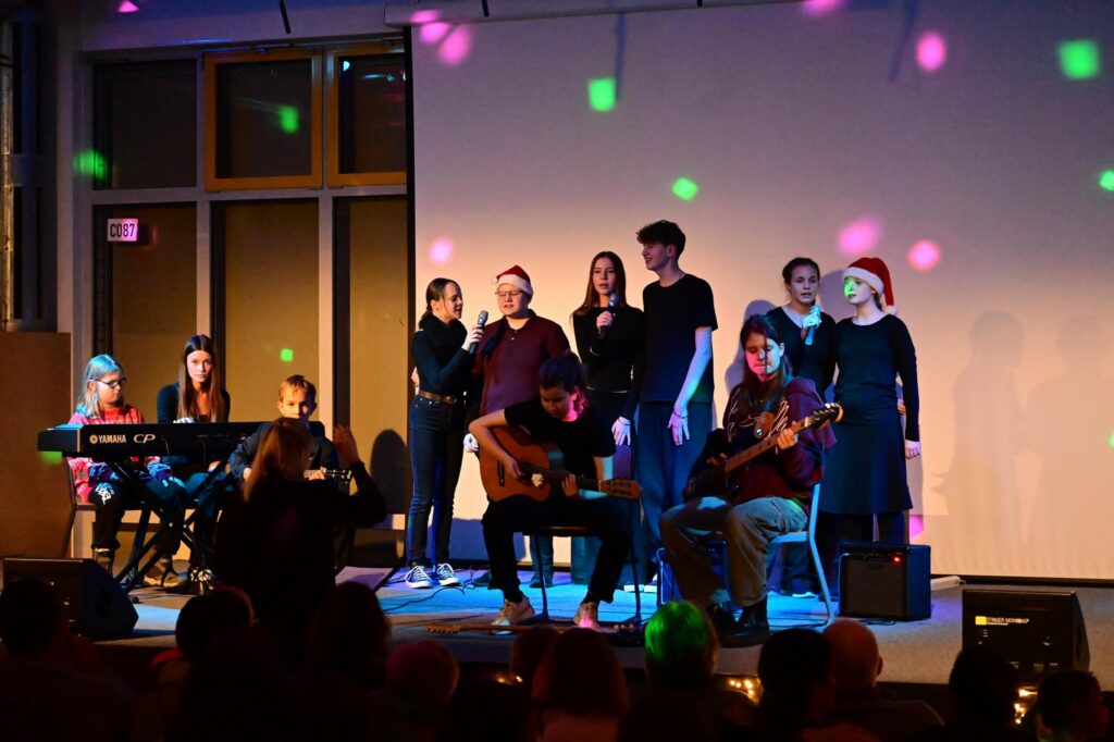 Band-AG der Schule auf der Bühne mit Gitarren, Bass, Keyboard und Sänger:innen, Publikum im Vordergrund, farbiges Bühnenlicht.