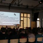 Blick von hinten auf das sitzende Publikum in der Mensa; auf der Bühne ist eine Projektionsfläche mit Hinweisen für die Vorleser:innen zu sehen.