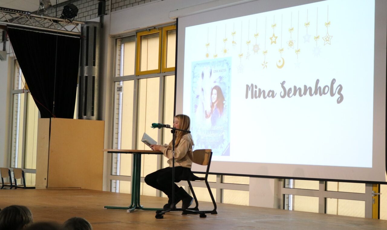 Schülerin liest an einem Stehtisch aus einem Buch; auf der Leinwand ist „Pferdeflüsterer Mädchen – Rubys Entscheidung“ zu sehen.