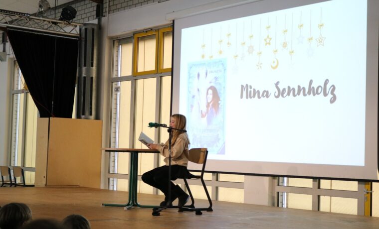 Schülerin liest an einem Stehtisch aus einem Buch; auf der Leinwand ist „Pferdeflüsterer Mädchen – Rubys Entscheidung“ zu sehen.
