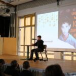 Schüler sitzt auf der Bühne und liest; auf der Leinwand ist das Buch „Und wenn ich zurückhaue?“ zu erkennen.