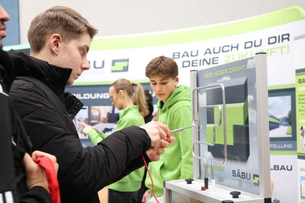 Ein Schüler testet an einem Mitmach-Modell handwerkliches Geschick, während weitere Jugendliche aufmerksam zusehen. Der Fokus liegt auf praktischem Ausprobieren und gemeinsamer Erfahrung.