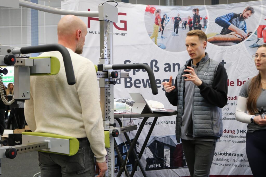 Ein Aussteller erklärt an einem Trainingsgerät Abläufe aus dem Gesundheits- und Bewegungsbereich. Die Szene zeigt intensive Beratung und fachliche Gespräche auf Augenhöhe.