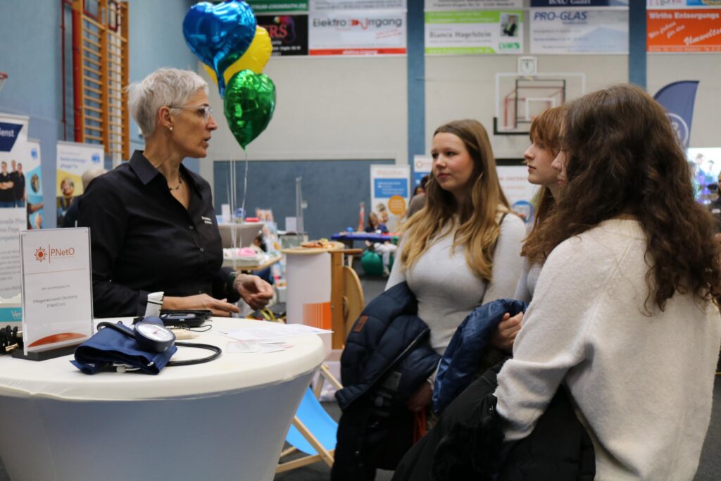 Drei Schülerinnen führen ein Gespräch mit einer Ausstellerin aus dem Pflegebereich. Die ruhige Gesprächssituation hebt die Bedeutung individueller Beratung hervor.